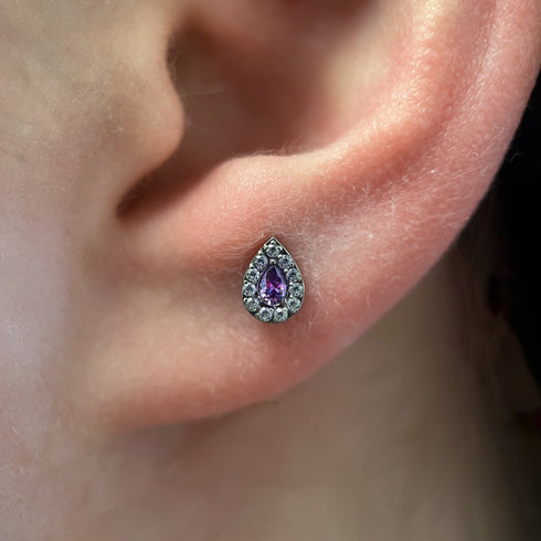 Close-up of an ear wearing a teardrop-shaped piercing with a purple gemstone.